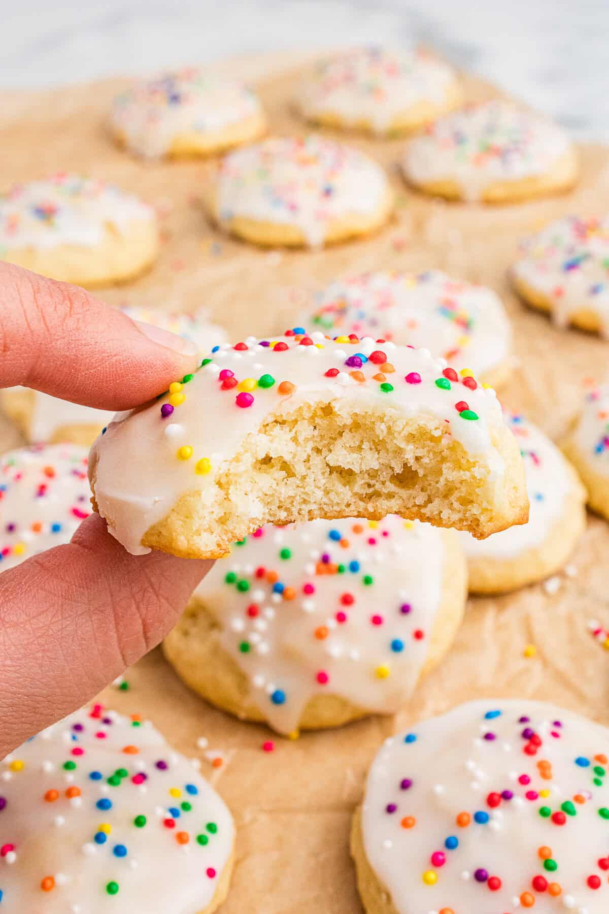 A hand holds a half-eaten Italian ricotta cookie with white icing and colorful sprinkles, revealing its soft, fluffy interior. More cookies are scattered in the background on parchment paper, highlighting the festive and homemade presentation.