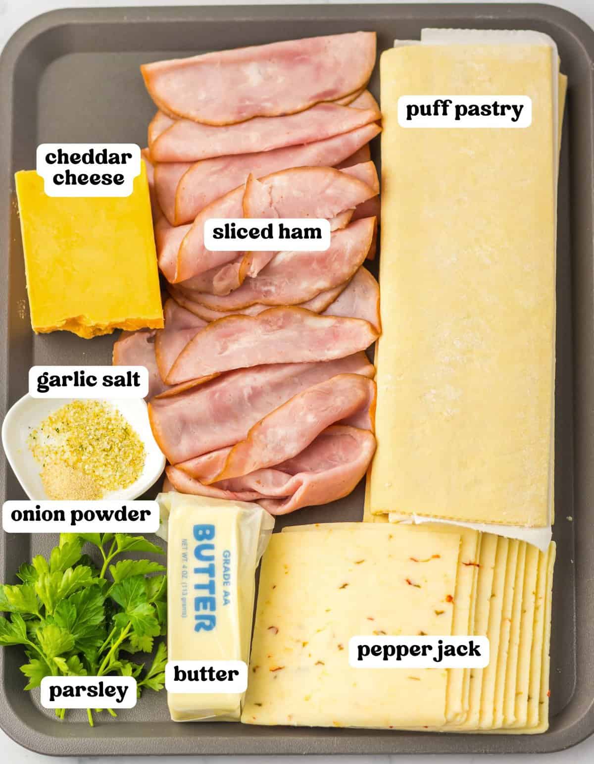 Tray with ingredients for puff pastry Christmas trees: sliced ham, cheddar cheese, pepper jack cheese, puff pastry sheets, butter, parsley, garlic salt, and onion powder.
