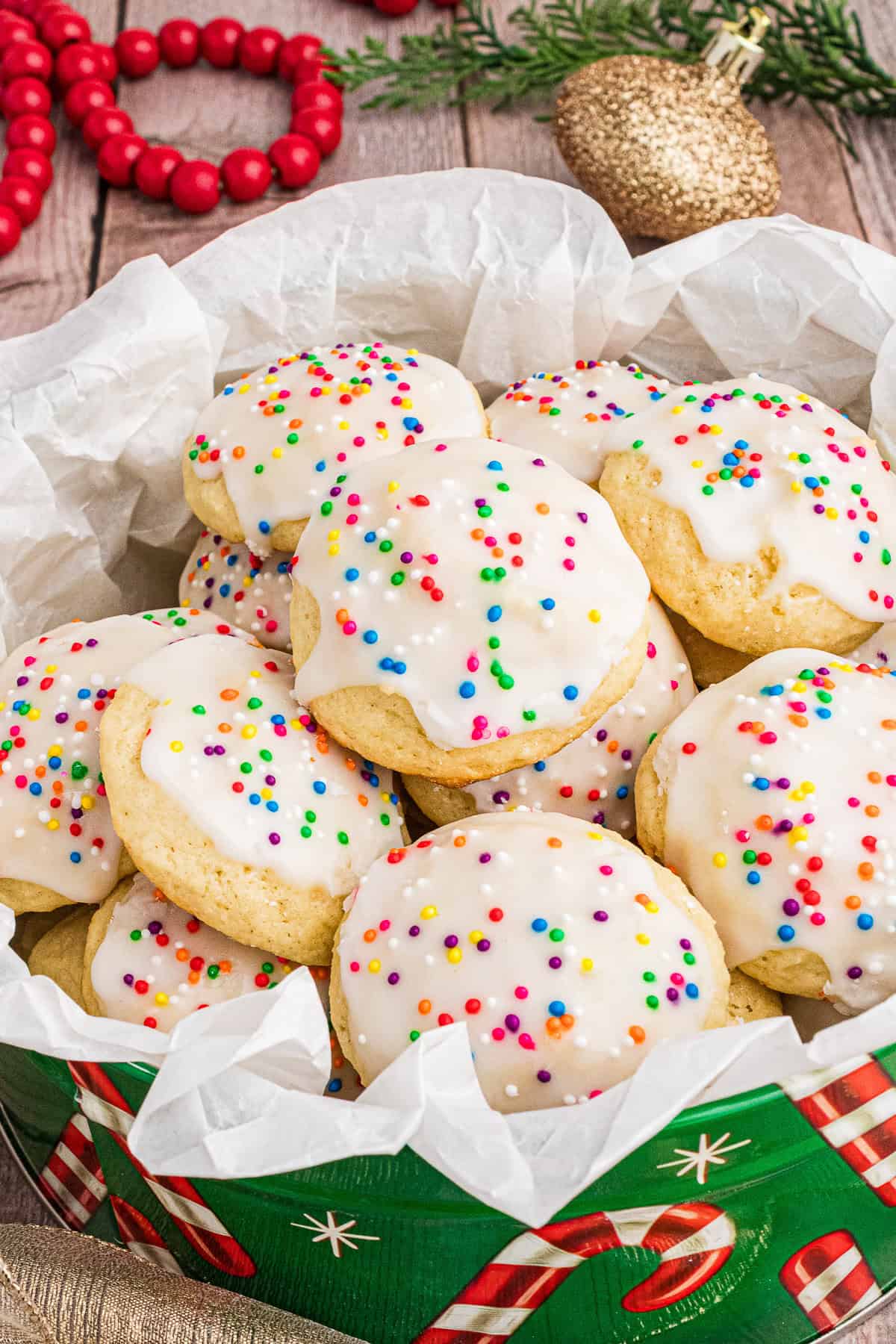 A festive display of Italian ricotta cookies with white icing and colorful sprinkles, piled in a holiday-themed tin lined with parchment paper. The background features red wooden beads, a gold ornament, and green pine branches, enhancing the Christmas setting.