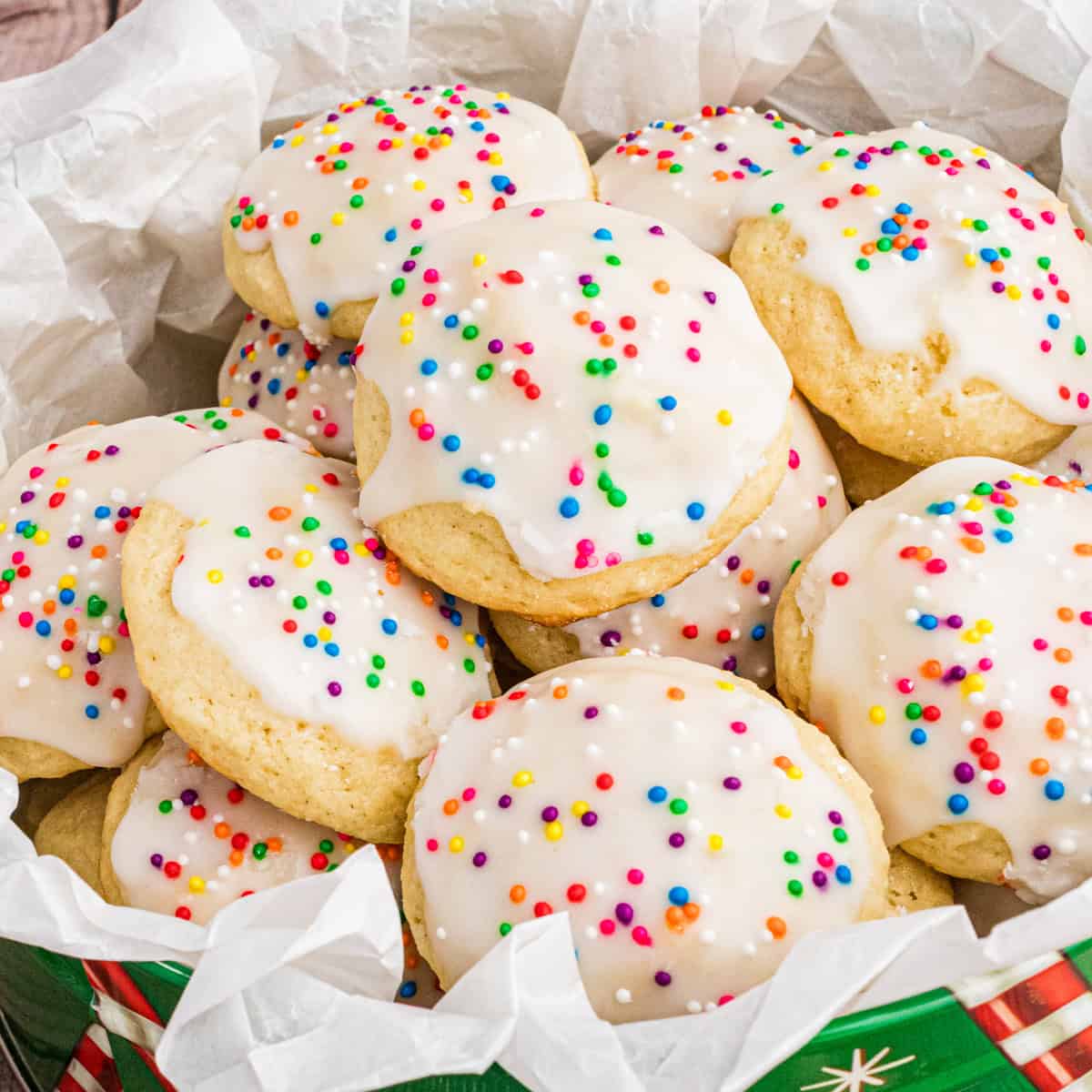 A close-up of Italian ricotta cookies topped with smooth white icing and colorful round sprinkles, arranged in a festive green tin lined with parchment paper. The cookies are soft, fluffy, and stacked to showcase their homemade charm.