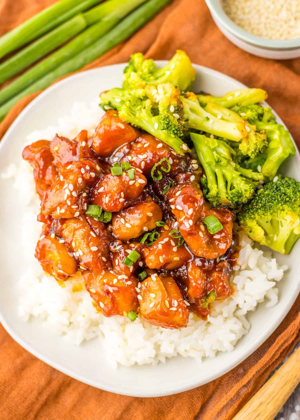 A plate of mall food court style bourbon chicken with sesame seeds and green onions, served with steamed broccoli on a bed of white rice.