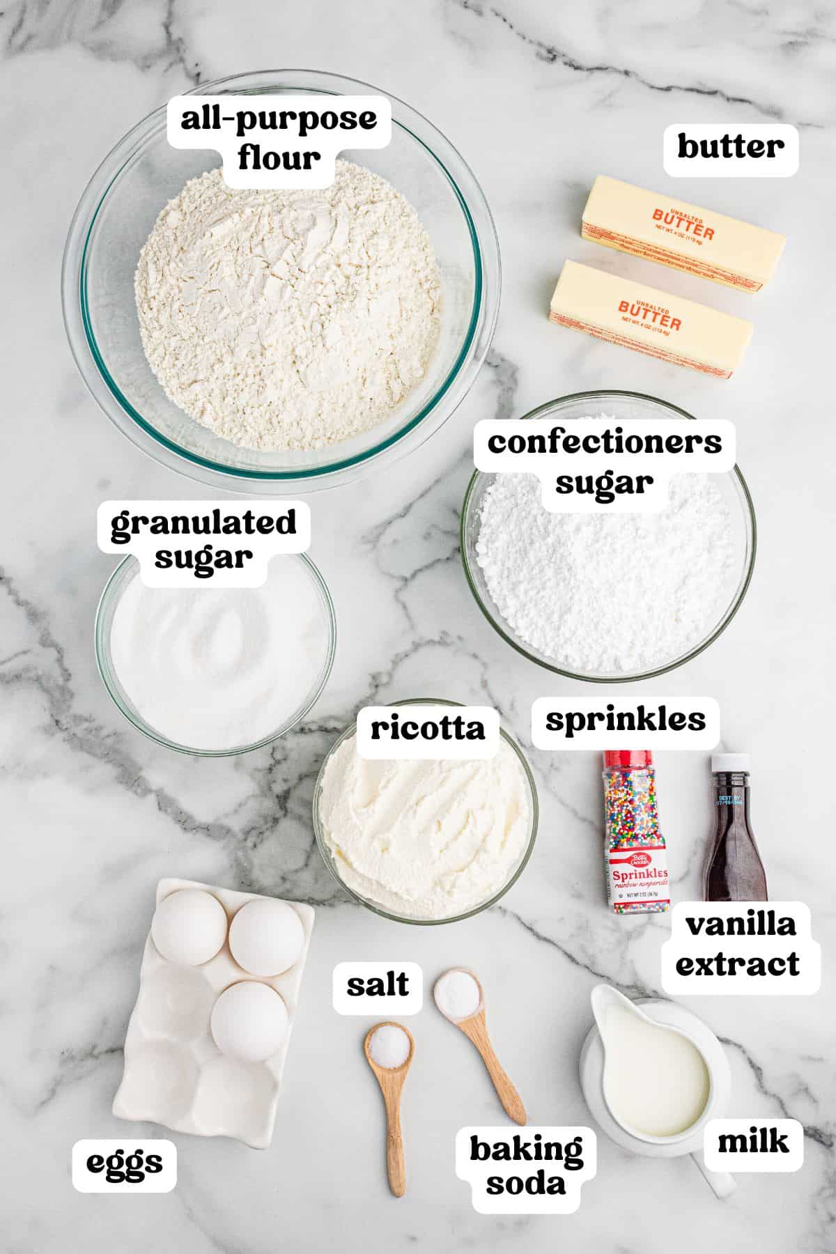 A top-down view of ricotta cookie ingredients labeled with text. The image includes bowls of all-purpose flour, granulated sugar, confectioners sugar, and ricotta, along with sticks of butter, eggs, salt, baking soda, milk, vanilla extract, and colorful sprinkles, all neatly arranged on a marble countertop.