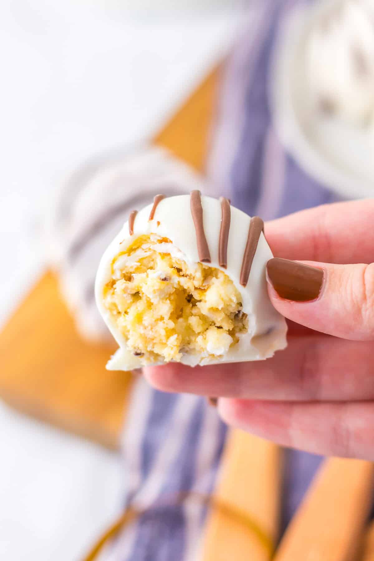 A hand holding a white chocolate-coated truffle (Zebra Cake Truffle) with a bite taken, showing the yelllow cream cheese and cake filling inside.